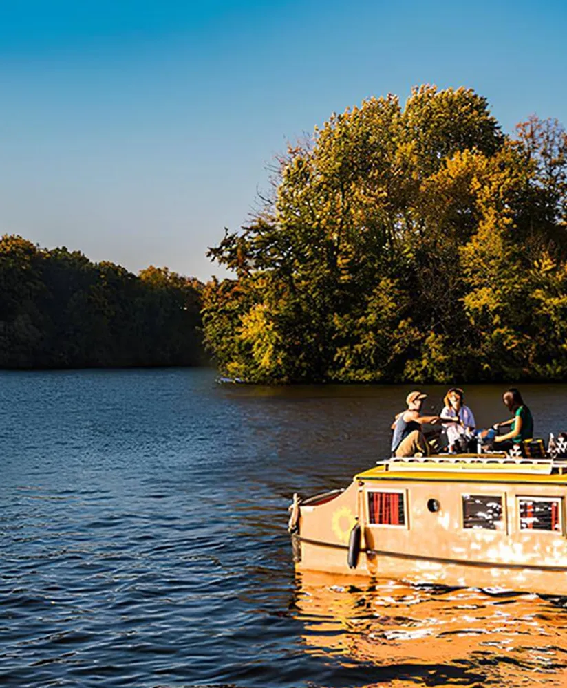 Kompaktes Hausboot auf ruhiger Spree wird von der untergehenden Sonne angestrahlt mit 3 Menschen auf dem Dack beim Abendessen.