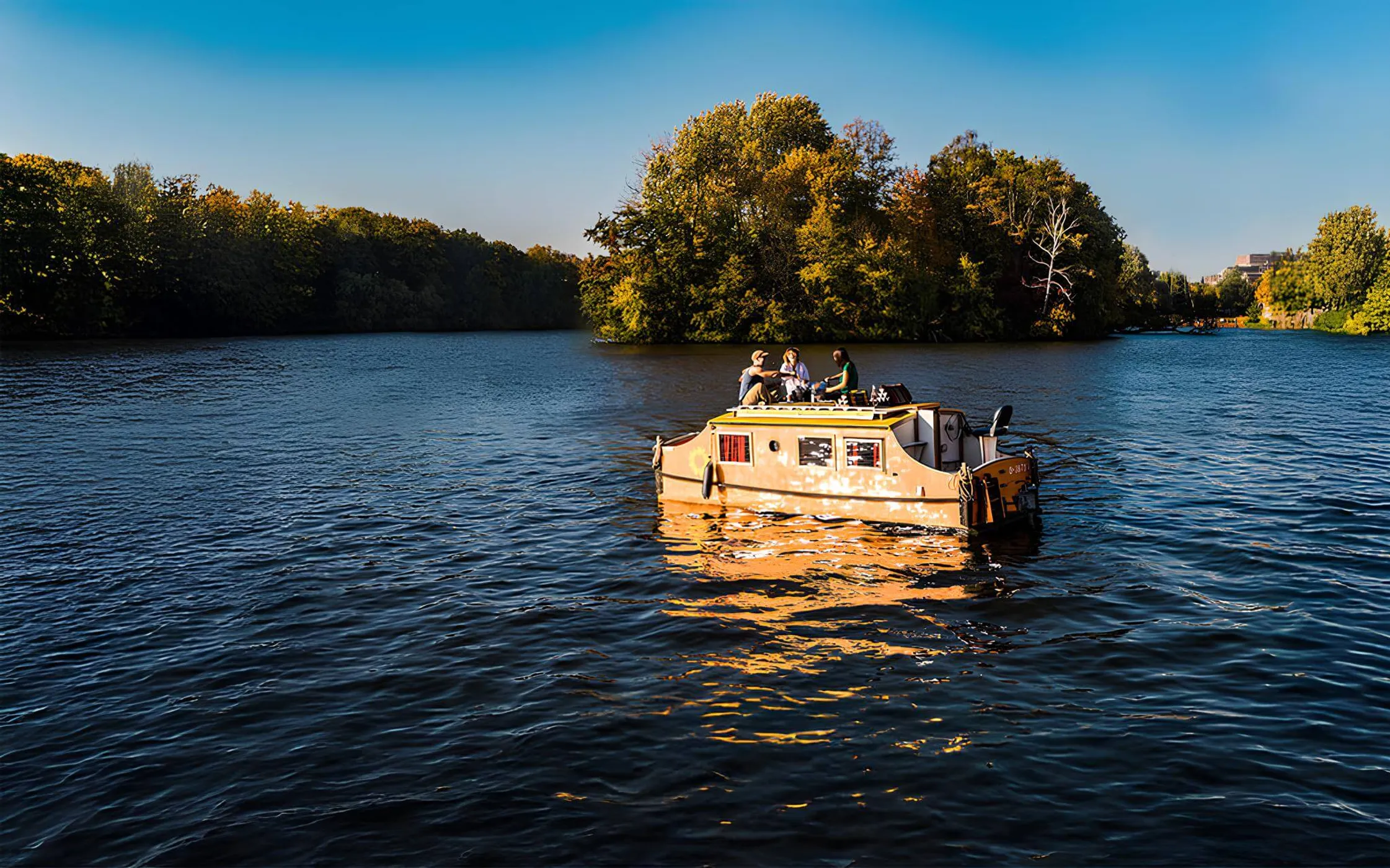 Kompaktes Hausboot auf ruhiger Spree wird von der untergehenden Sonne angestrahlt mit 3 Menschen auf dem Dack beim Abendessen.